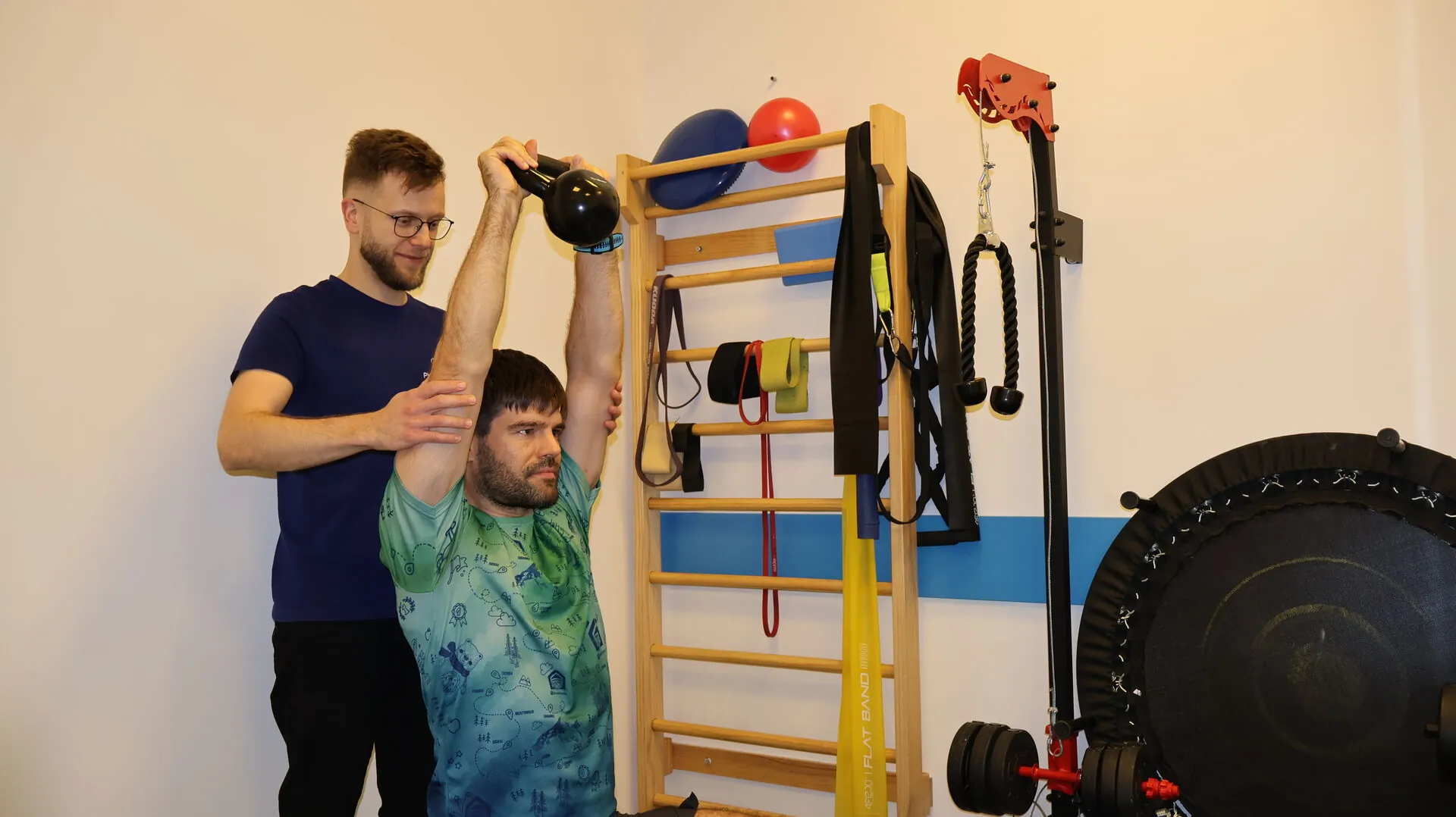 Fizjoterapeuta asystujący pacjentowi przy wyciskaniu kettlebell nad głowę w gabinecie Physiomedica